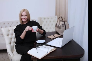 Genç bir kadın oturuyor, gülümsüyor ve bir fincan kahveyle masada çalışıyor. Tanımlanamayan fotoğraf. İş sabahı ya da iş gezisi kavramı. Boşluğun kopyası.