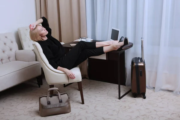A tired business woman rests with her feet on the table. A room in the hotel . A suitcase and a travel bag stand next to each other. A woman at work on a business trip. The view from the top . A copy
