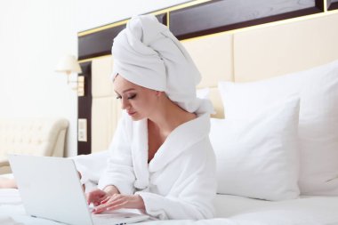 Yatakta bornozlu ve kafasında havlu olan genç bir kız dizüstü bilgisayarla çalışıyor. Uzaktan öğrenme ya da uzaktan çalışma kavramı. Ev ortamı. Boşluğun kopyası.