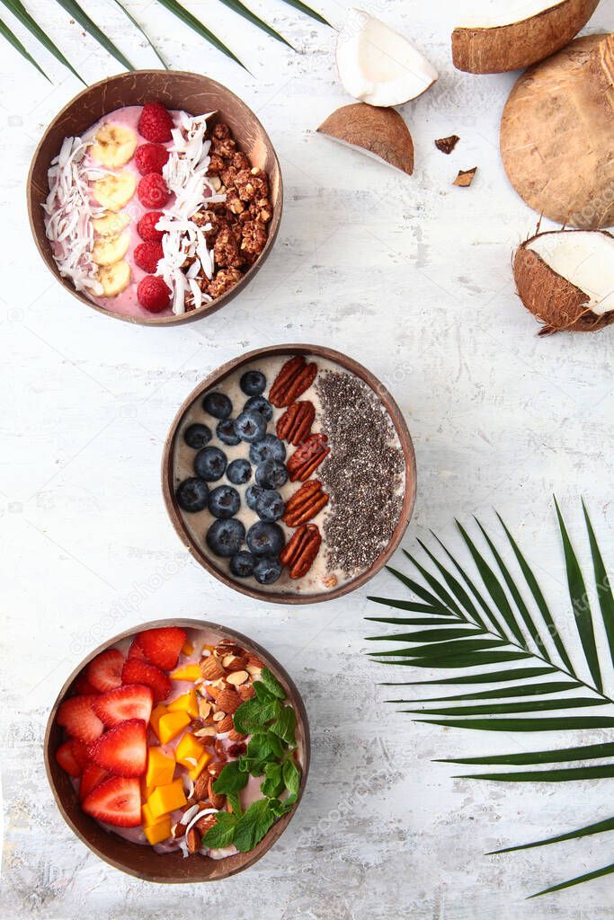 Balinese-style breakfasts. Muesli with yogurt and berries in plates made of coconut skin. Tropical tree leaves and coconuts. Light textured background. Wholesome breakfast. Top view. Copy space.