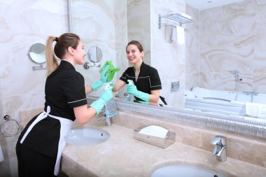 Temizlik üniformalı genç bir kız banyodaki aynayı temizliyor. Odaklanamıyor. Banyo mermer fayanslarla donatılmış. Pahalı bir otelde temizlik. Boşluğu kopyala.