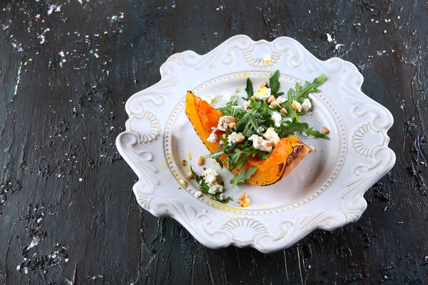 Запечённая тыква с сыром фета, с рукколой и кедровыми орехами на белой тарелке. Фото на темном текстурированном фоне. Вид сверху. Копирование пространства
.