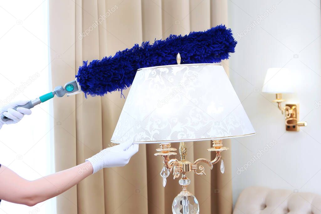 The maid's white-gloved hands with a long brush sweep the dust from the floor lamp. Unrecognizable photo, cleaning in a hotel or at home. Copy space.