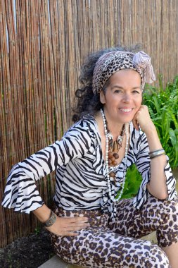 Dışarda oturan, egzotik giyinmiş, gülümseyen Creole kadını. Bambu çitler ve arka planda bitkiler. Açık renk.