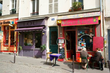 Montmartre Mahallesi 'nde renkli restoran ve butik cepheleri. İnsanlar dar bir kaldırımda bir masada oturuyor. Öğle yemeği saati, güneş ışığı. Paris, Fransa.                         