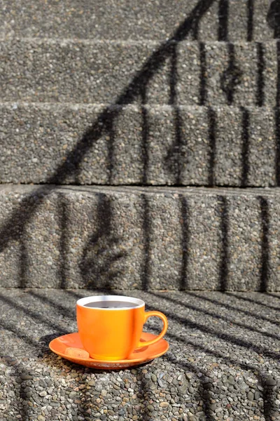 An orange colored cup and saucer are placed on a step of a grey stone staircase. Cup filled with coffee or hot chocolate. A little cookie is on the saucer. The shadow of the handrail draws the steam out of the cup. Sunlight.