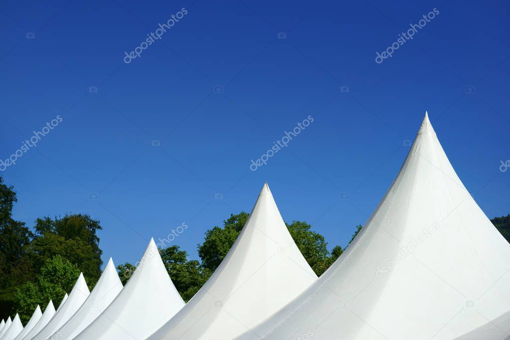 A dozen of white canopy top tents in a row. View on the points of the tents. Trees and a blue sky in the background. Copy space.