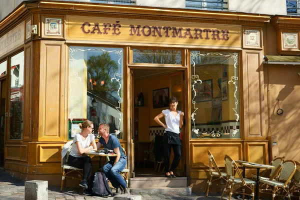 Montmartre Mahallesi 'ndeki bir kafenin ön cephesi ve terası. Bir bistro masasında oturan gülümseyen bir çift. Kapıda durup Nisan güneşini alan garson. Paris, Fransa.                      