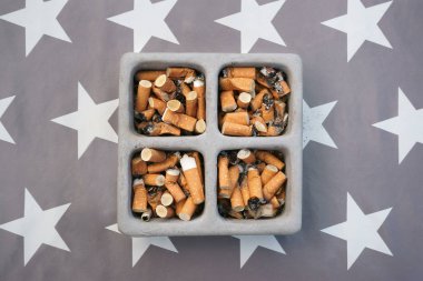 Sigara dolu bir kül tablası beyaz yıldızlı gri bir masa örtüsüne konmuş. Masaüstü görünümü.