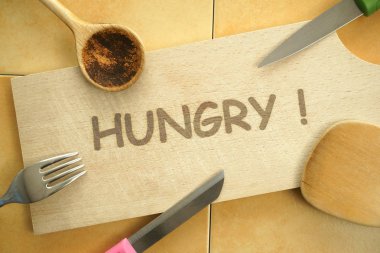 Acıktım! Tahta kesme tahtasındaki yazı. Bıçaklar, bir çatal, tahta kaşık ve tahtanın etrafında duran spatula. Masa üstü görüntüsü. Arkaplanda seramik fayanslar. 