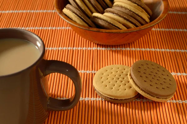 2つのチョコレートで満たされたクッキー ミルクティーと茶色のマグカップとクッキーでいっぱいの竹のボウル すべてのテラコッタオレンジ竹のテーブルセットに配置されます クローズアップ サイドライティング — ストック写真