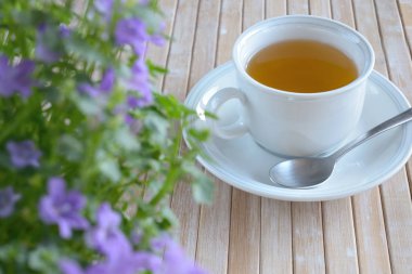 Bir fincan çay, çay tabağı ve kaşığıyla. Bulanık mor çan çiçekleri. Bambu masasına yerleştirilmiş eşyalar.