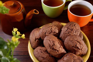 Sarı bir tabakta ev yapımı çikolatalı kurabiyeler, çay dolu iki renkli fincan, kahverengi çaydanlık ve koyu kahverengi ahşap bir masaya yerleştirilmiş sarı çiçekli bitki..
