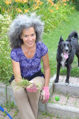 Bahçesinde yabani otları topladıktan ve Border Collie köpeğiyle yakalamaca oynadıktan sonra dinlenen gülümseyen olgun bir kadın. Arka planda zambaklar. 