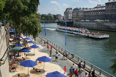 Seine nehrinin kıyısında şemsiyeli kumsallar. Turistlerle dolu bir Bateaux-Mouches gezi teknesi. Arcole Köprüsü ve arka planda Ile Saint-Louis. Çiçekçi kutuları solda. Ile de la Cite sağda. Paris, Fransa.