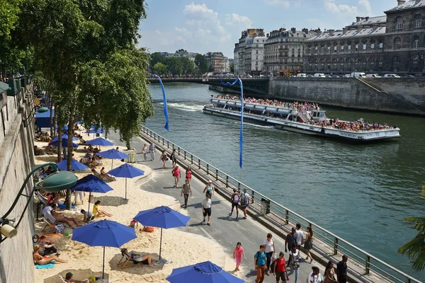 Seine nehrinin kıyısında şemsiyeli kumsallar. Turistlerle dolu bir Bateaux-Mouches gezi teknesi. Arcole Köprüsü ve arka planda Ile Saint-Louis. Çiçekçi kutuları solda. Ile de la Cite sağda. Paris, Fransa.
