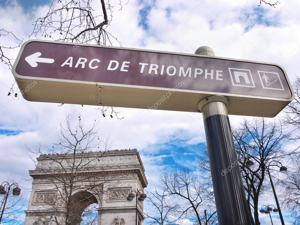 The Arc de Triomphe and its street signal with an arrow indicating the way how to access it. Sky background and winter trees. Place Charles de Gaulle, Paris, France. 