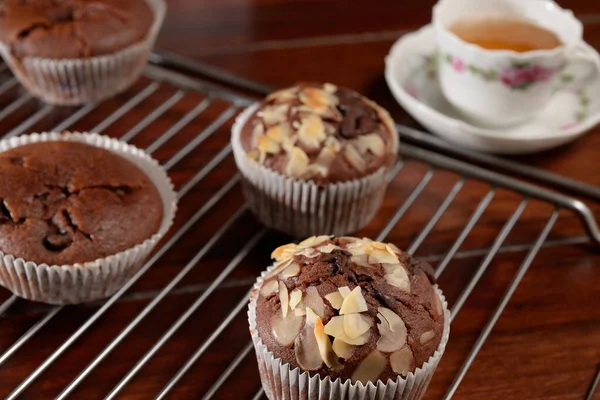 冷却ラックに自家製チョコレートマフィン 奥にはお茶が入った磁器のカップ プレーンチョコレートマフィンとアプリコットジャムとスライスしたアーモンドといくつか 暗い木製のテーブルの上に置かれたすべての項目 — ストック写真