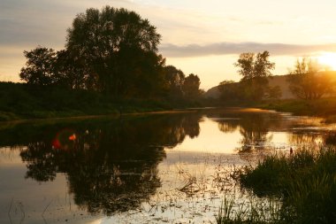 Sakin bir nehrin üzerinde günbatımının manzarası ve sonbaharın başlarında sahilde ağaçlar.