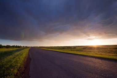Gün batımında ufka giden yaz manzarası yolu ve mavi gökyüzünde dramatik bulutlar.