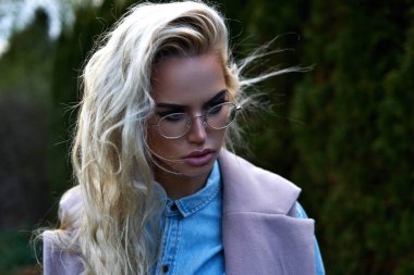 Sarışın, güzel bir kız. Kışlık kıyafetleriyle dışarıda dolaşıyor. Arka plan olarak thuja 'yı kullanıyor ve kameraya poz veriyor.