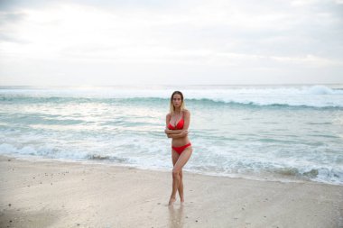 Güzel seksi bir manken beyaz kumsalda, kırmızı bikinili, bronzlaşmış bir mayo, uzun sarı saçlar, muhteşem göğüsler ve dolgun dudaklar, Bali 'de sağlıklı okyanus dalgaları.