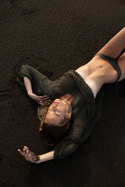 Güzel seksi manken siyah bir mayoyla siyah kumsalda uzanıyor. Uzun sarı saçlı, muhteşem göğüsleri ve dolgun dudakları var. Bali adasında sağlıklı okyanus dalgaları bulutları.