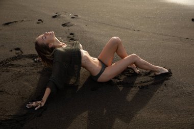 Güzel seksi manken siyah bir mayoyla siyah kumsalda uzanıyor. Uzun sarı saçlı, muhteşem göğüsleri ve dolgun dudakları var. Bali adasında sağlıklı okyanus dalgaları bulutları.
