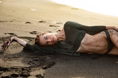 Güzel seksi manken siyah bir mayoyla siyah kumsalda uzanıyor. Uzun sarı saçlı, muhteşem göğüsleri ve dolgun dudakları var. Bali adasında sağlıklı okyanus dalgaları bulutları.