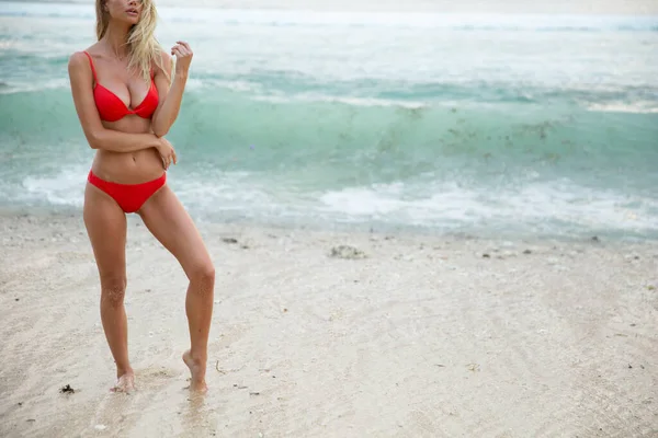 一位漂亮性感的女模特穿着红色比基尼泳衣 有着一头晒黑的身躯 一头金色的秀发 丰满的胸部和丰满的嘴唇 在雪白的沙滩上悠闲自在 — 图库照片