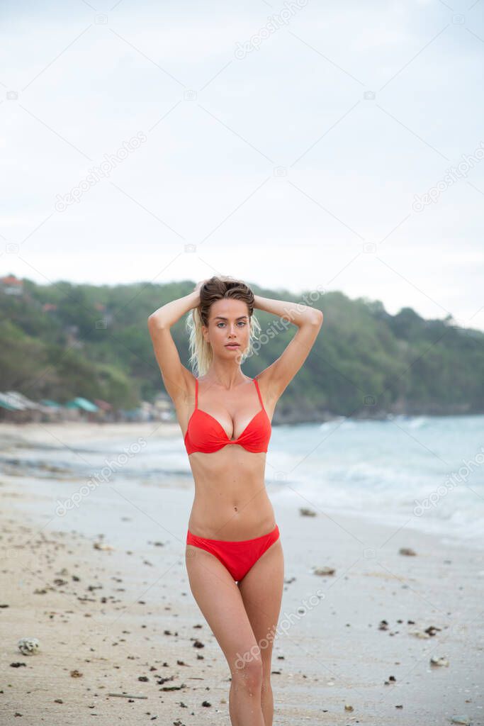 A beautiful sexy girl model relax on white sand beach in red bikini swimsuit with a tanned body long blonde hair magnificent breasts and puffy lips Healthy nature ocean waves clouds on Bali