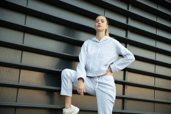 Şehir merkezinde poz veren atletik bir kız modelin hafif bir eşofmanı var. Kaputu, spor ayakkabıları. Sokakta atış stili..
