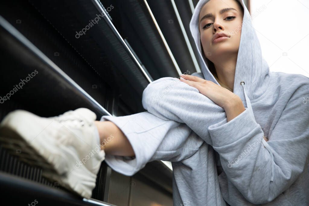 Athletic girl posing in an urban city The model has a light tracksuit hood sneakers Stylish sports shooting on the street A beautiful athletic body face skin lips A girl with a menacing look.