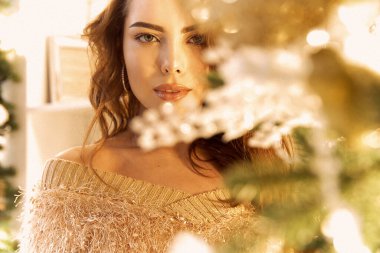 Güzel şirin kız hediyesini açar, hediyeleri kucaklar, gülümser ve bir Noel ağacını yeni yıl süslemeleriyle süsler. Yeni yıl makyajı, saç stili ve bronz ten..
