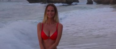 Güzel, seksi kız gün batımında kumsalda dalgalarla eğleniyor ve gülümsüyor. Parlak, sıcak güneş ve kumsalda beyaz kumlar var. Okyanustaki bazı sörfçüler dalgalarını alıyor.