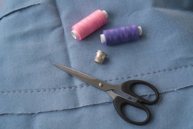 Eteğin bir kısmını dikiş, makas, yüksük ve iki makara iplikle kes. Mavi kumaşın üzerinde iki adet pembe ve mor iplik, metal kapak ve makas var.