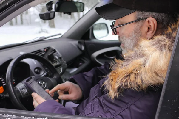 A driver is sitting in the car and looks at the mobile phone. Cold weather, snow-covered trees in the winter forest. Winter travel.