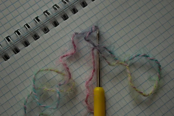 Metal crochet hook with a yellow tip, multi-colored thread on a notebook with a spring. Merino yarn. Threads for knitting and crocheting. Creativity and Hobbies.