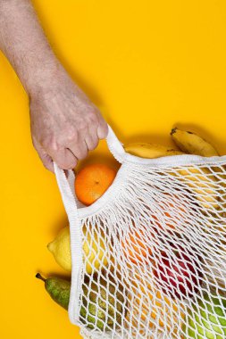 A human hand holds a string bag with fresh fruits on yellow paper background