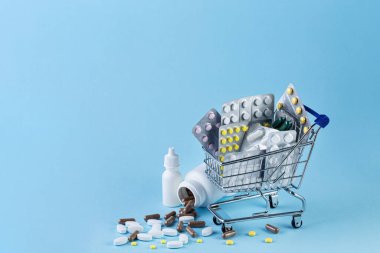 Medicine in shop trolley and pills spilling out of white bottle on blue background
