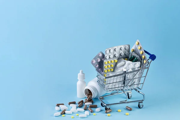 Medicine in shop trolley and pills spilling out of white bottle on blue background — 스톡 사진