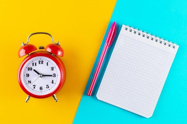 Alarm saatini mavi ve sarı arka planda bir kalem ve defterin yanına koy. Okulda ve üniversitede okuyor. Zaman yönetimi konsepti, iş planlaması. Üst Manzara Düz Yat
