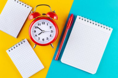 Alarm saatini mavi ve sarı arka planda bir kalem ve defterin yanına koy. Okulda ve üniversitede okuyor. Zaman yönetimi konsepti, iş planlaması. Üst Manzara Düz Yat