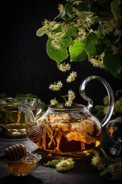 Çaydanlık ve bardak, ıhlamur çayı ve çiçeklerle. Koyu arka planda güzel uçan ıhlamur çiçekleri, yaratıcı çiçek düzeni. Bitki çayı.
