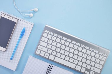 Asgari çalışma alanı. Çalışma masasının yaratıcı düz yatay fotoğrafı. Not defteri, klavye, kağıt not defteri ve mavi arkaplanlı malzemelerle üst kattaki ofis masasında. Ev fikrinden çalış..