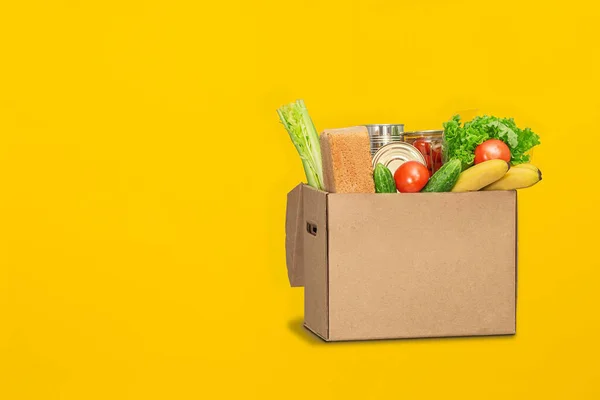 Donation box with food on a yellow background. Coronavirus food delivery. Volunteer brings a box of food. Donation, quarantine concept.  Volunteer collecting food. Copyspace.