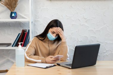Yüksek ateşi ve baş ağrısı olan koruyucu maske takan bir kadın karantina sırasında dizüstü bilgisayarla çalışıyor.,