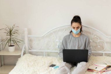 Korumacı tıbbi maske takan genç bir kadın öğrenciler için öğrenim görüyor. Karantinaya alın. Freelancer, evde çalış, online.