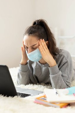 Yüksek ateşi ve baş ağrısı olan koruyucu maskeli bir kadın karantina sırasında dizüstü bilgisayarla çalışıyor.,