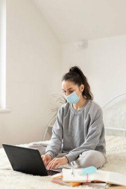 Korumacı tıbbi maske takan genç bir kadın öğrenciler için öğrenim görüyor. Karantinaya alın. Freelancer, evde çalış, online.